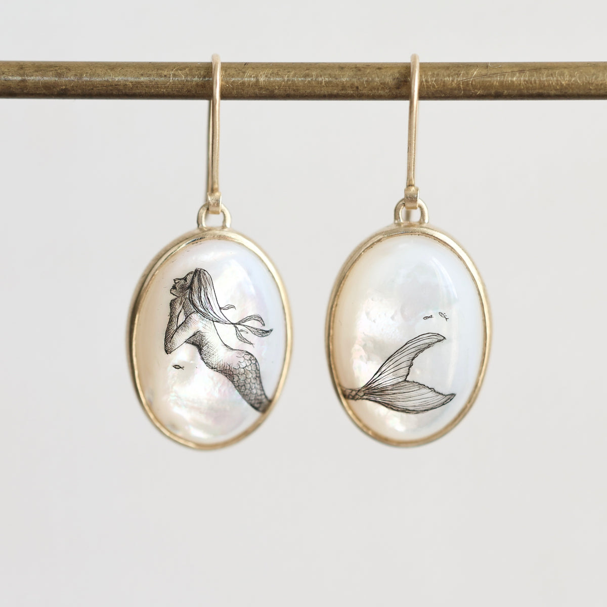 A pair of gold-tone oval drop earrings with mother-of-pearl cabochons featuring black ink drawings of mermaids, hanging from a gold rod against a light gray background.