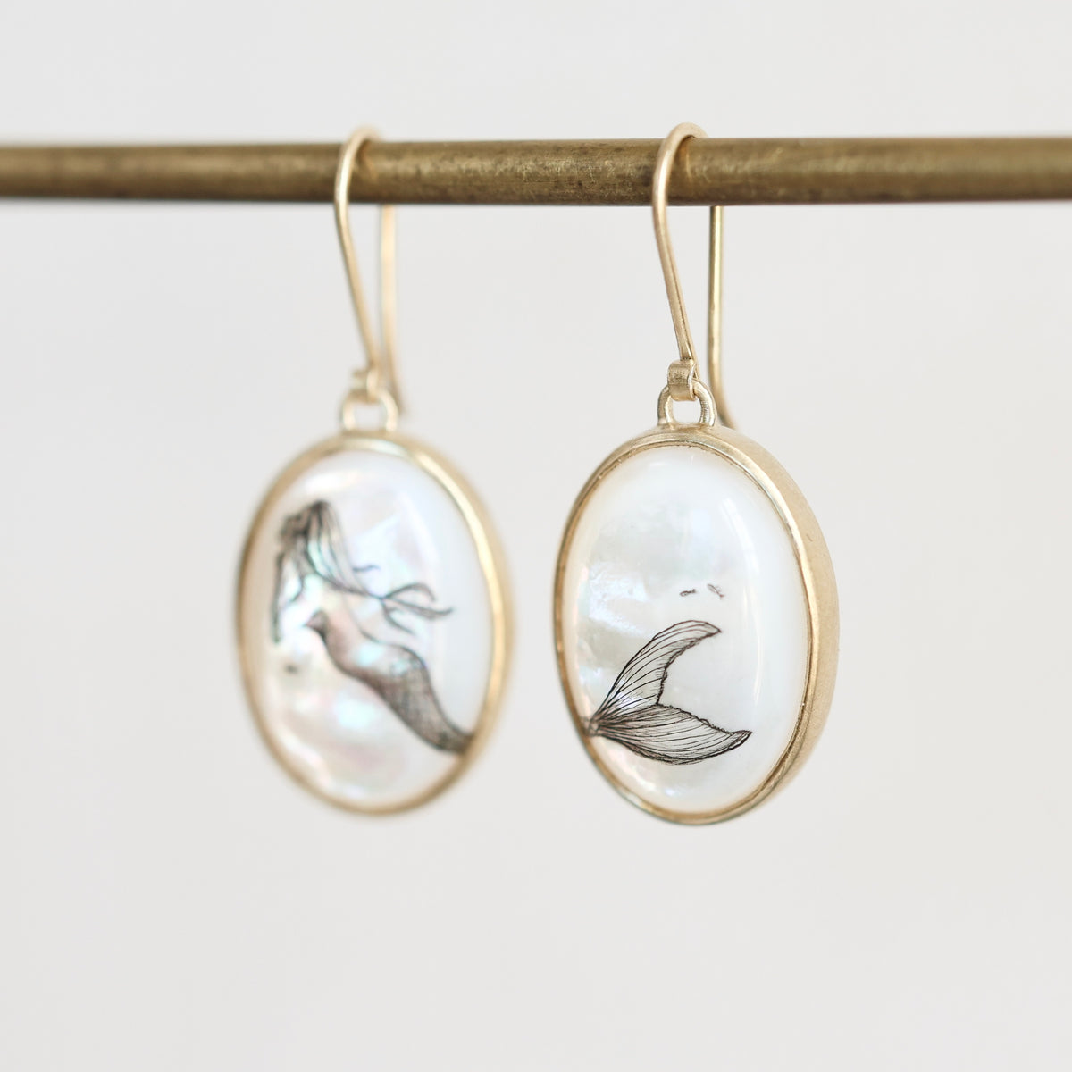 A pair of gold-tone oval drop earrings with white mother-of-pearl inlays featuring black mermaid tail illustrations, hanging from a gold rod against a light gray background.