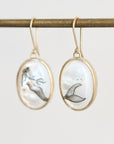 A pair of gold-tone oval drop earrings with white mother-of-pearl inlays featuring black mermaid tail illustrations, hanging from a gold rod against a light gray background.