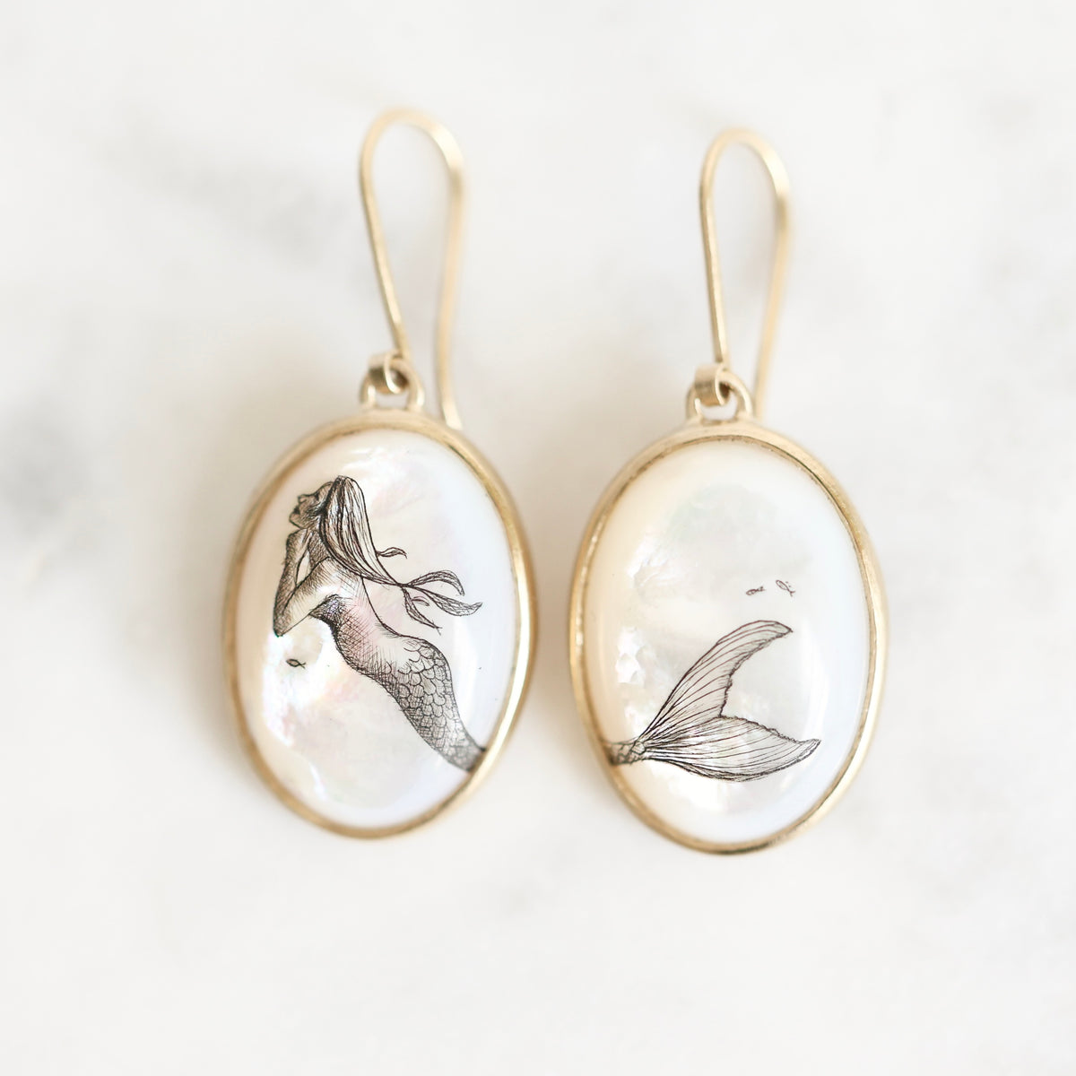 A pair of gold drop earrings with oval white mother-of-pearl cabochons featuring black line art illustrations of a mermaid and a fish tail, displayed on a light marble background.