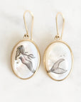 A pair of gold drop earrings with oval white mother-of-pearl cabochons featuring black line art illustrations of a mermaid and a fish tail, displayed on a light marble background.