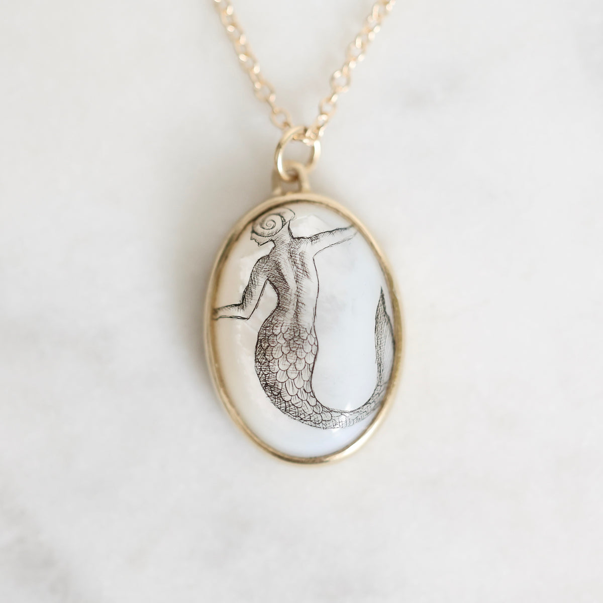 Gold oval pendant necklace featuring a mother-of-pearl inlay with a hand-painted mermaid design, shown on a light marble background.