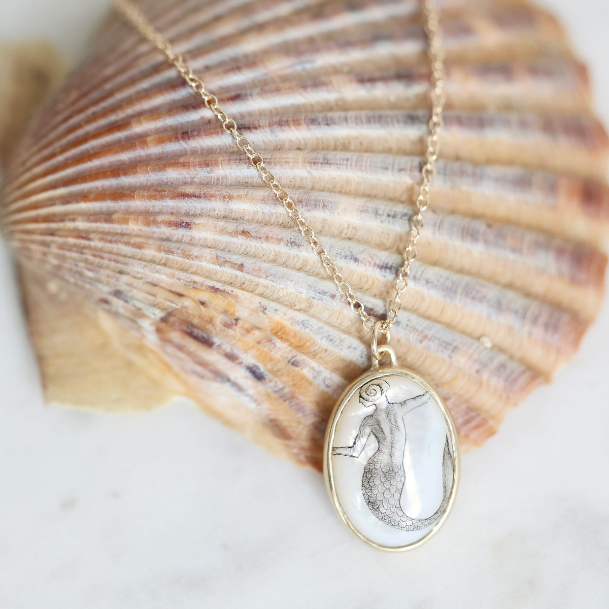 Gold oval pendant necklace featuring a mermaid design, displayed on a large seashell against a light background.