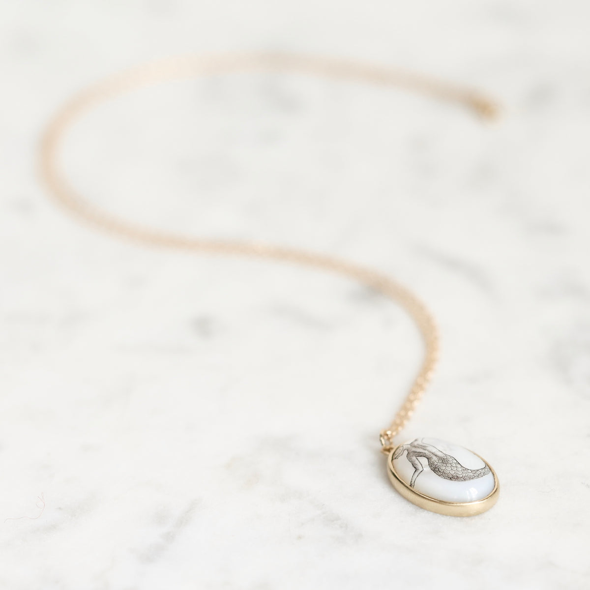 A delicate gold chain necklace with an oval pendant featuring a white background and a gray sketch of a high-heeled shoe, displayed on a light marble surface.