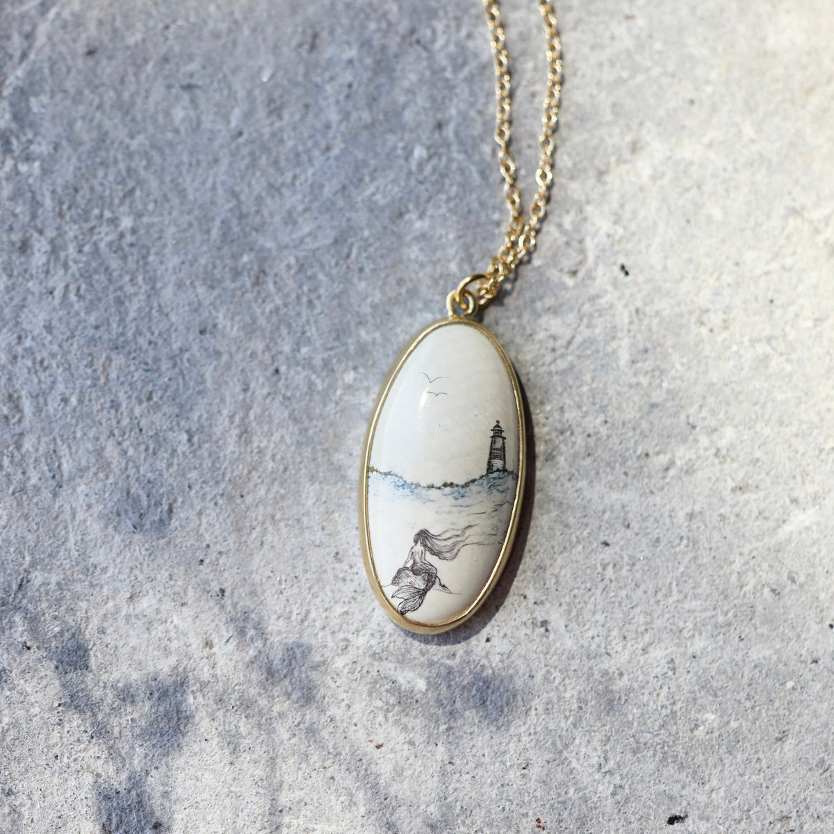 An oval pendant necklace with a gold-toned frame and chain, featuring a hand-painted scenic illustration of a lighthouse and a woman on a white background, resting on a textured gray stone surface.