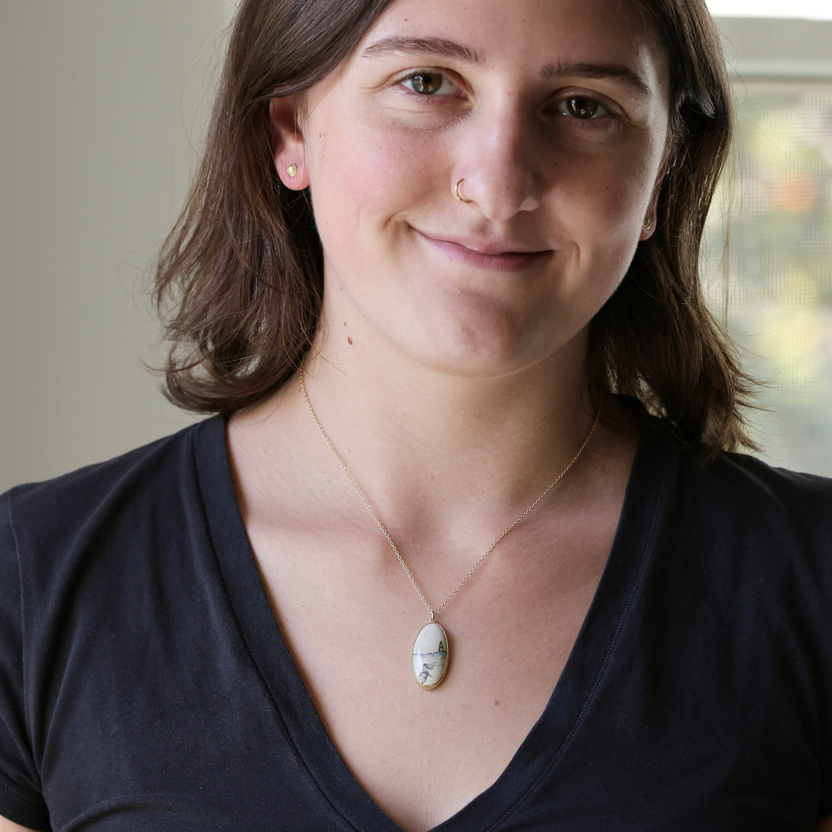 A woman with brown hair wearing a black V-neck top and a delicate gold chain necklace with a small white oval pendant, photographed in a softly lit indoor setting.