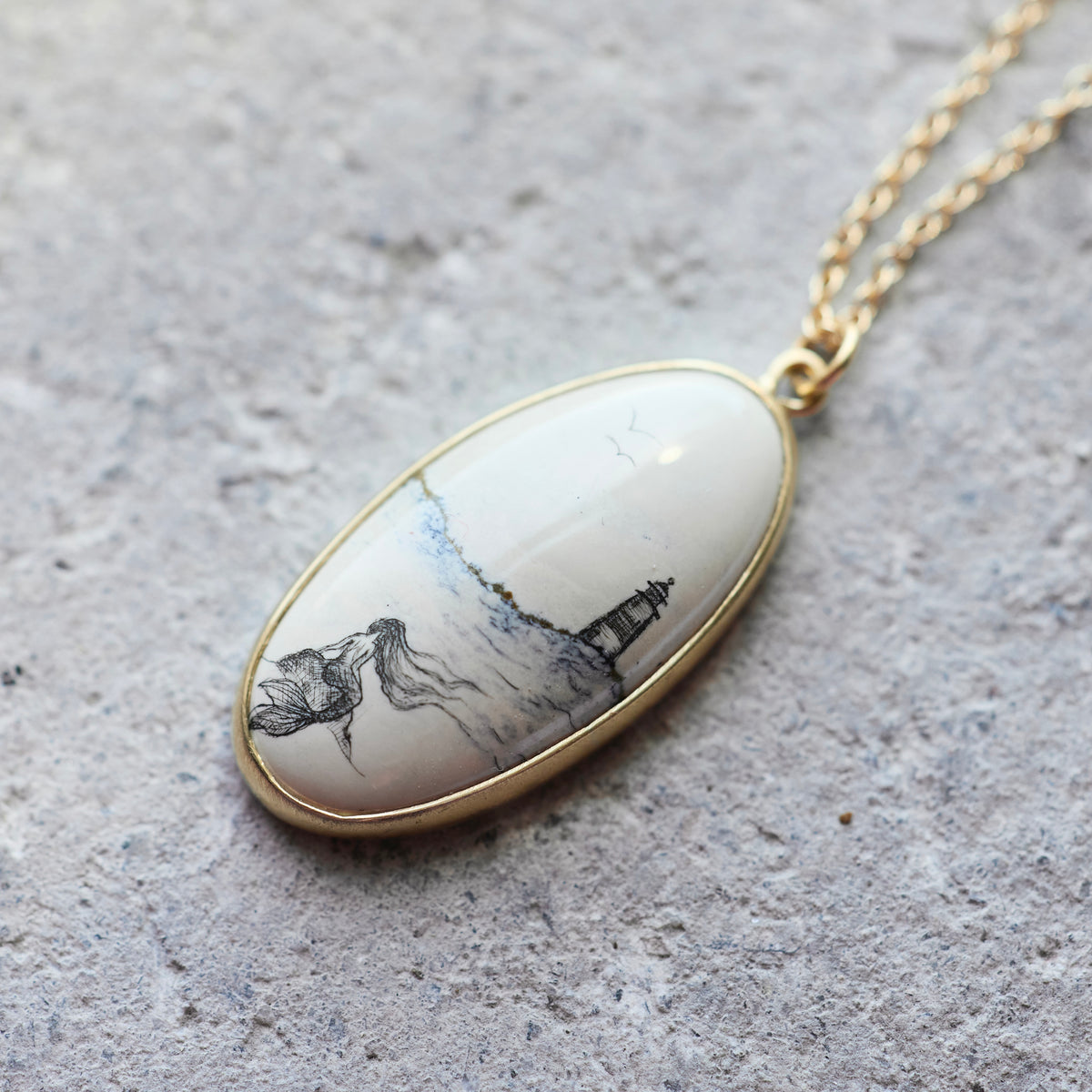 An oval pendant necklace with a gold-tone border and chain, featuring a cream-colored stone or resin inlay printed with a black ink-style landscape of a lighthouse and trees, resting on a textured gray stone surface.
