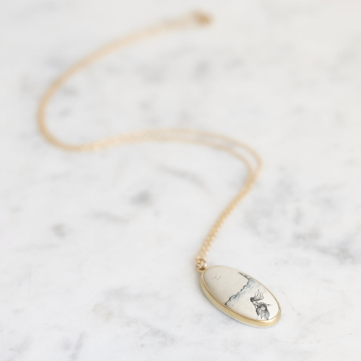 A gold chain necklace with an oval pendant featuring a white stone with black and blue veining, displayed on a light gray marble surface.