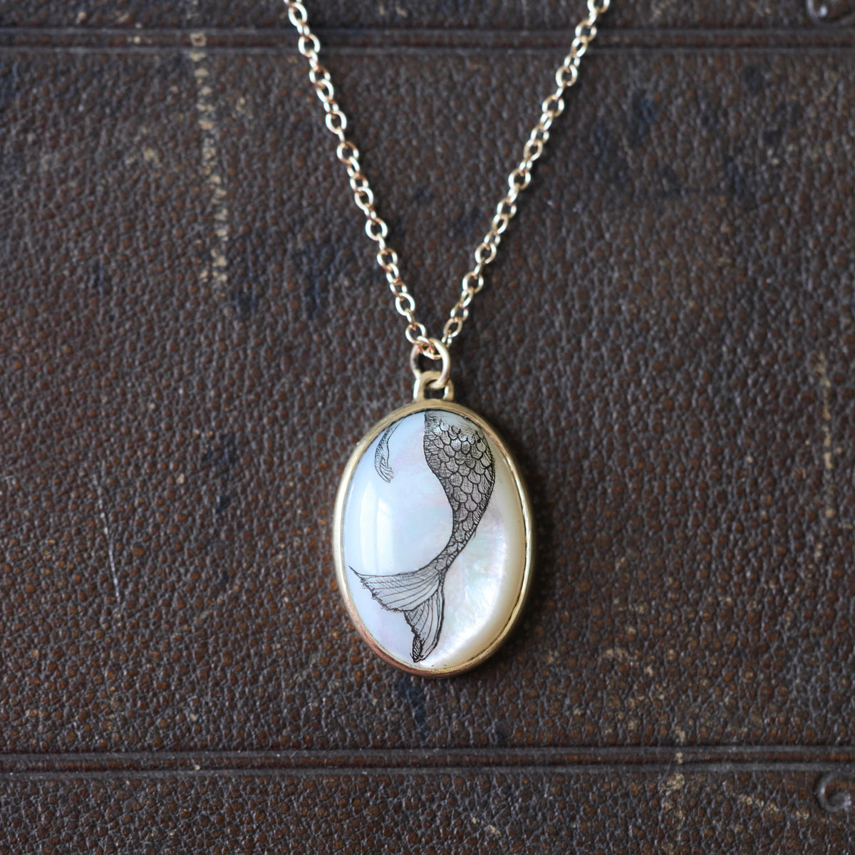 An oval pendant necklace featuring a black ink mermaid illustration on a white mother-of-pearl background, set in a gold-tone bezel with a silver-tone chain, displayed on a dark brown textured leather surface.