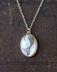 An oval pendant necklace featuring a black ink mermaid illustration on a white mother-of-pearl background, set in a gold-tone bezel with a silver-tone chain, displayed on a dark brown textured leather surface.