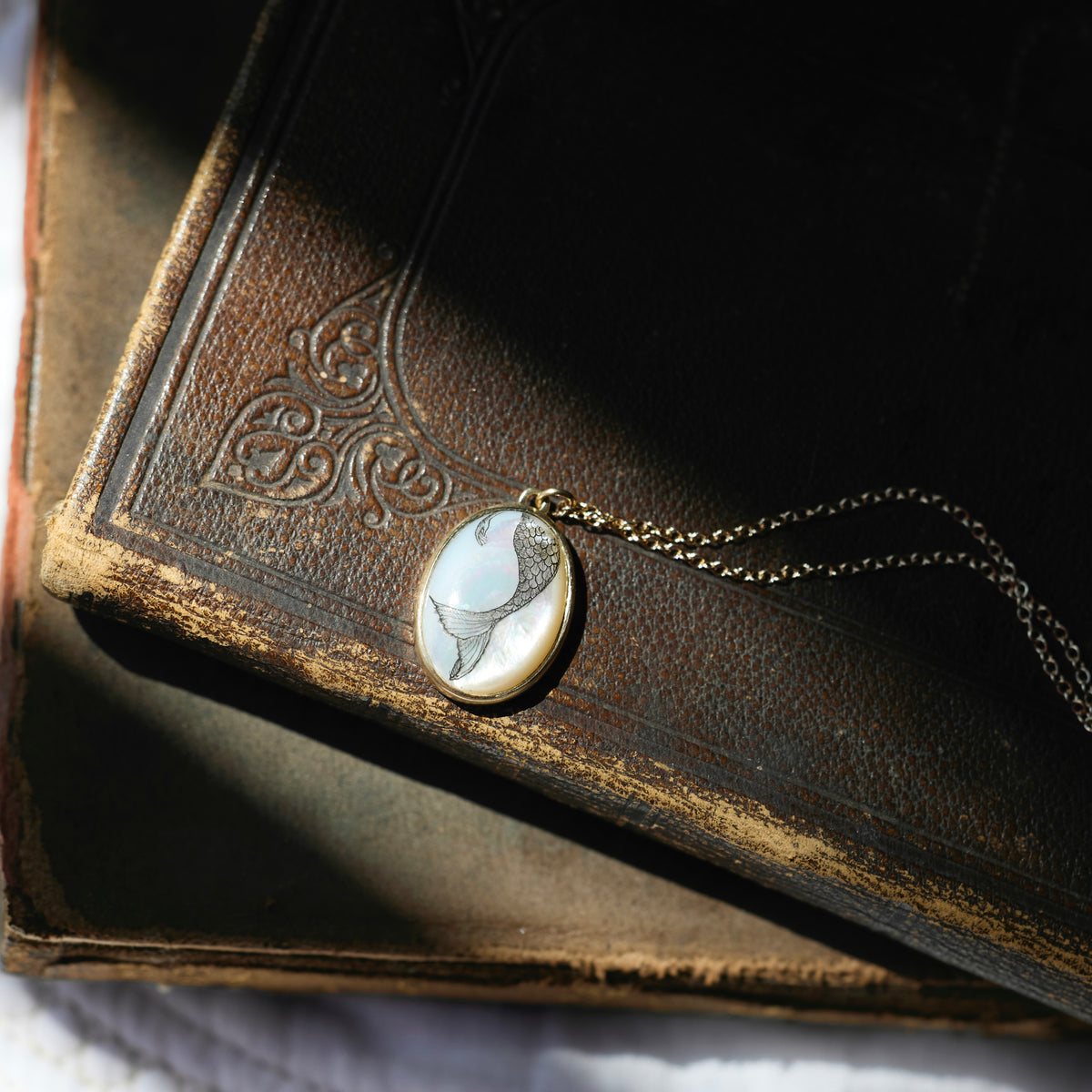 A close-up of a vintage oval pendant necklace with a white and gray bird design set in gold, resting on an antique brown leather-bound book.