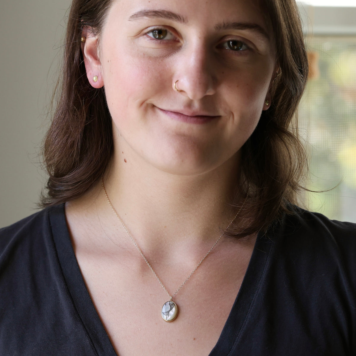 A woman with brown hair wearing a delicate gold chain necklace with a small oval white shell pendant, photographed in a close-up portrait.