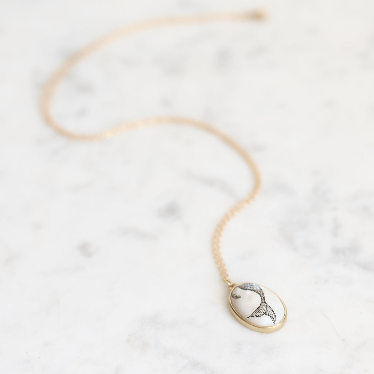 A delicate gold chain necklace with an oval pendant featuring a black and white fish design, displayed on a light gray marble surface.