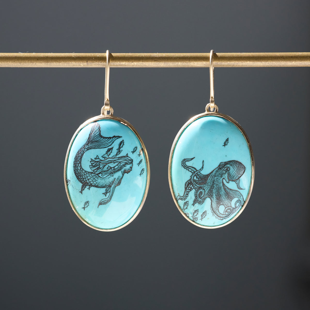 A pair of oval drop earrings hanging from a gold-toned rod against a dark gray background. Each earring has a light aqua blue stone featuring a black mermaid or octopus illustration, set in a gold-tone frame.