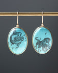 A pair of oval drop earrings hanging from a gold-toned rod against a dark gray background. Each earring has a light aqua blue stone featuring a black mermaid or octopus illustration, set in a gold-tone frame.