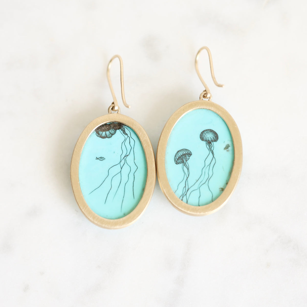 A pair of oval drop earrings with gold-tone frames and hooks, featuring a light blue turquoise engraved with black jellyfish illustrations, displayed on a white marble surface.