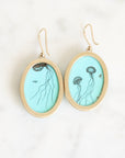 A pair of oval drop earrings with gold-tone frames and hooks, featuring a light blue turquoise engraved with black jellyfish illustrations, displayed on a white marble surface.