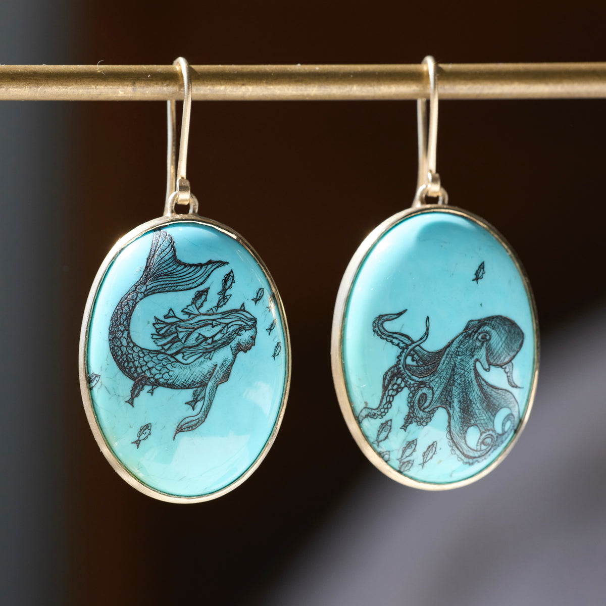 A pair of oval drop earrings with light blue turquoise-style cabochons, one featuring a black mermaid illustration and the other an octopus, hanging from gold-tone hooks on a gold rod against a dark blurred background.