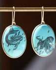 A pair of oval drop earrings with light blue turquoise-style cabochons, one featuring a black mermaid illustration and the other an octopus, hanging from gold-tone hooks on a gold rod against a dark blurred background.