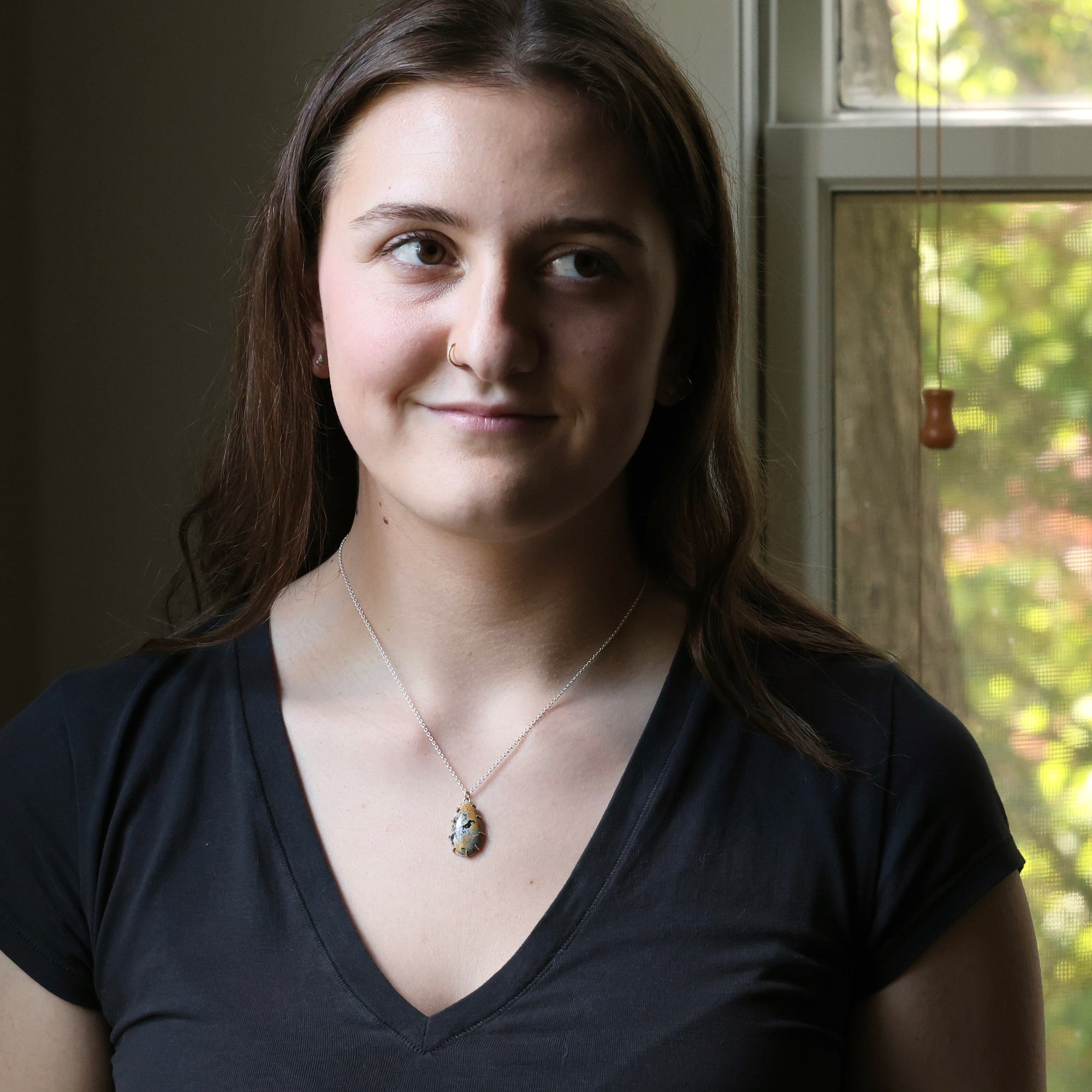 Woman wearing a black shirt with a necklace, standing in front of a window with greenery outside.