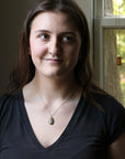Woman wearing a black shirt with a necklace, standing in front of a window with greenery outside.