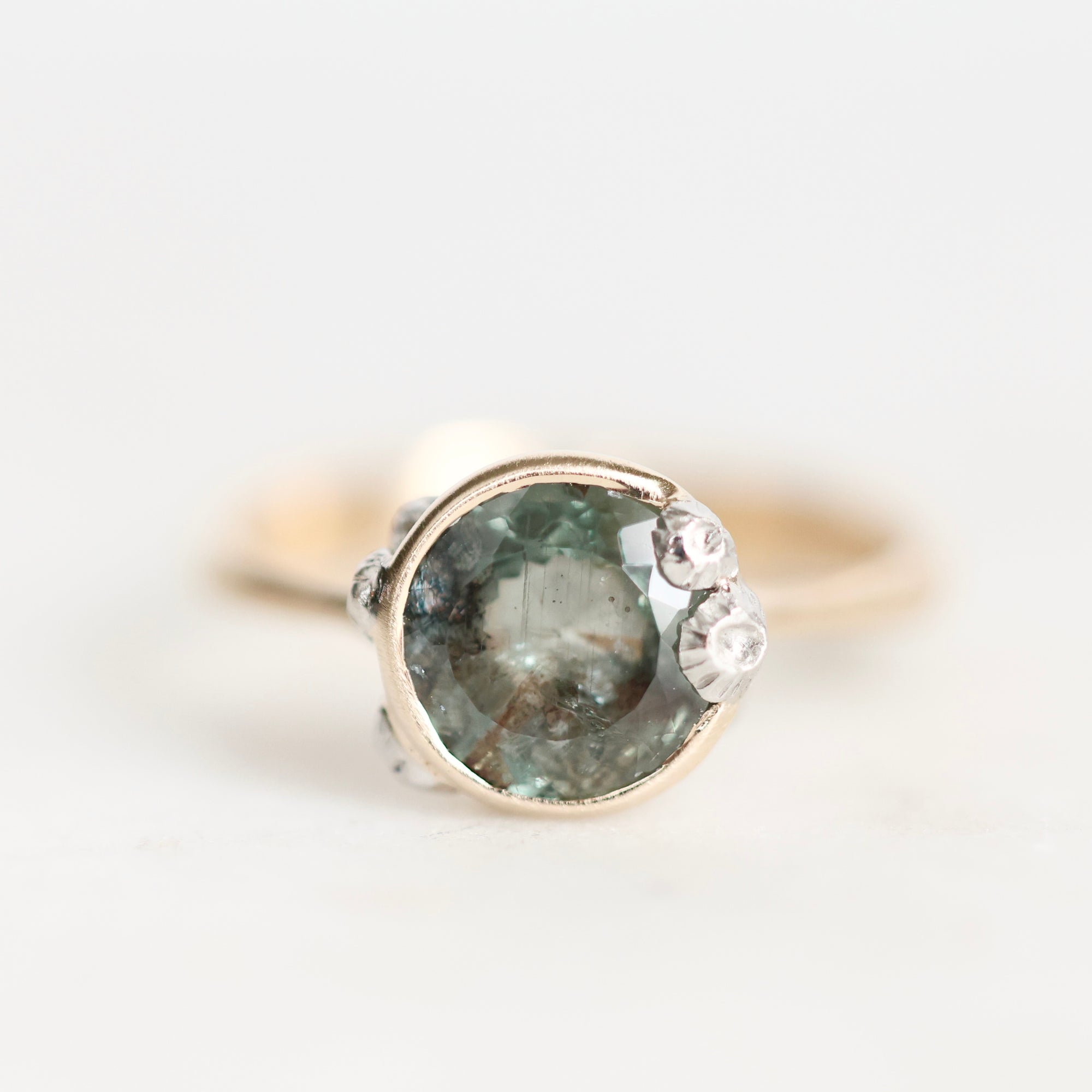A close-up shot of a ring featuring a round, faceted green gemstone in a silver-toned setting, accented with three small clear crystals on the right side, against a plain white background.