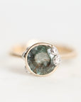 A close-up shot of a ring featuring a round, faceted green gemstone in a silver-toned setting, accented with three small clear crystals on the right side, against a plain white background.