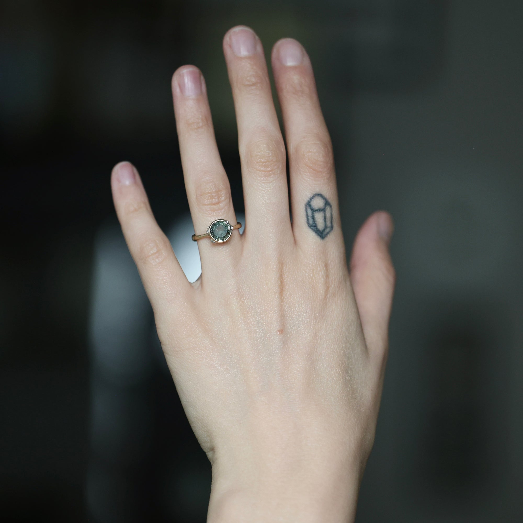 A close-up of a person's hand wearing a delicate ring with a small oval green stone and a rose gold band. The hand also features a small geometric tattoo on the middle finger, set against a dark, blurred background.