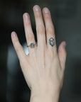 A close-up of a person's hand wearing a delicate ring with a small oval green stone and a rose gold band. The hand also features a small geometric tattoo on the middle finger, set against a dark, blurred background.