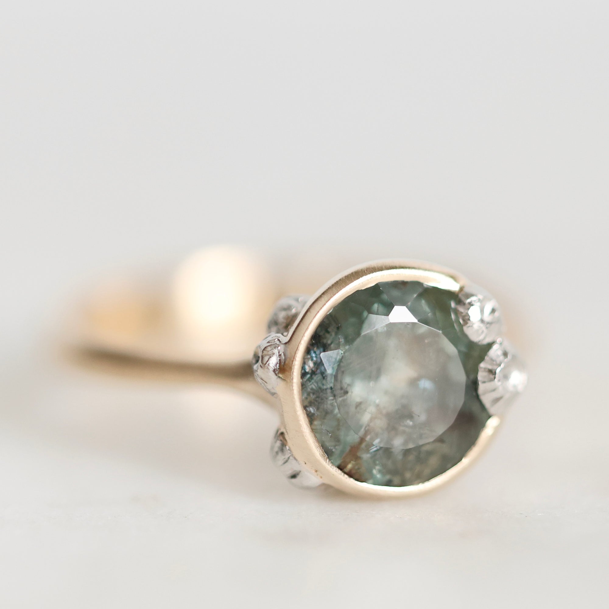 A close-up shot of a vintage-style ring featuring a large, round, pale green-gray gemstone in a gold bezel setting, accented by small clear stones on the shoulders, set against a soft, light gray background.
