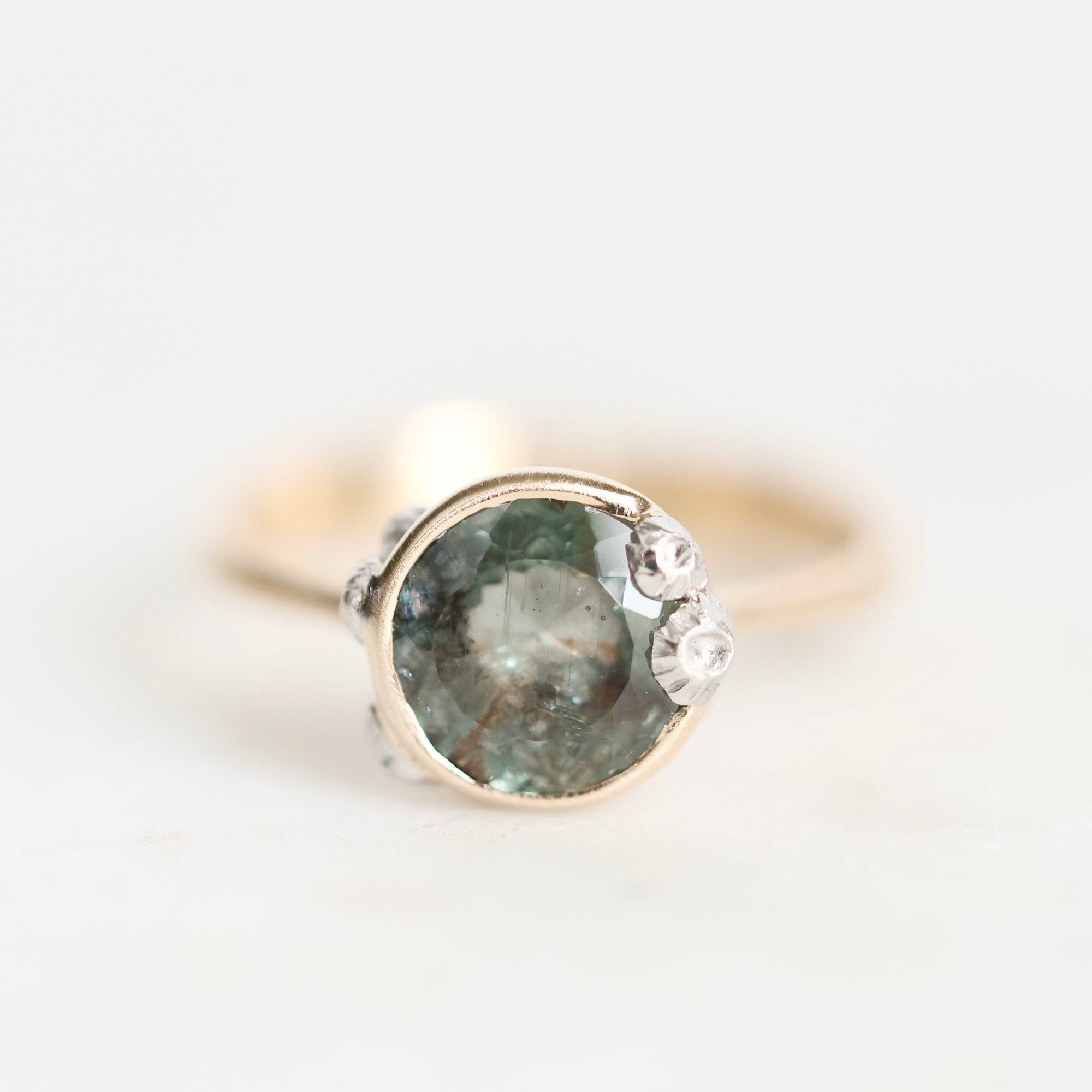 A close-up shot of a ring featuring a round green gemstone in a gold bezel setting, accented with three small clear crystals on the right side, set against a plain white background.