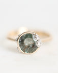 A close-up shot of a ring featuring a round green gemstone in a gold bezel setting, accented with three small clear crystals on the right side, set against a plain white background.