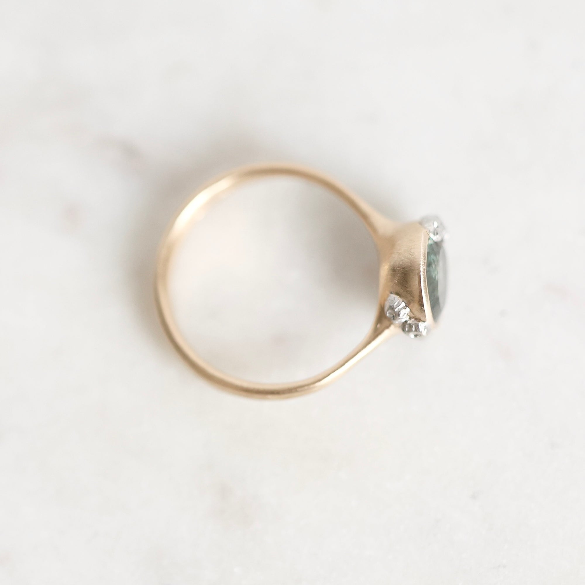 A delicate gold ring with a small oval green gemstone and two tiny clear accent stones, photographed on a light marble surface.