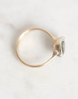 A delicate gold ring with a small oval green gemstone and two tiny clear accent stones, photographed on a light marble surface.
