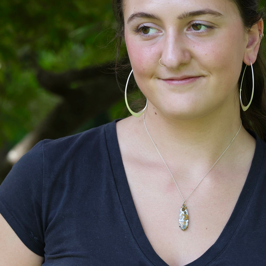 A woman wearing a silver-toned necklace with a small oval pendant and matching large hoop earrings, photographed outdoors with a blurred green background.