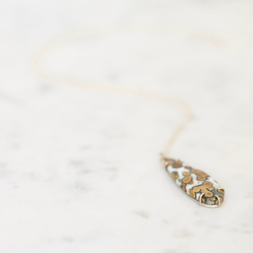A delicate gold necklace with a teardrop-shaped pendant featuring a white background and a gold floral pattern, resting on a light marble surface.
