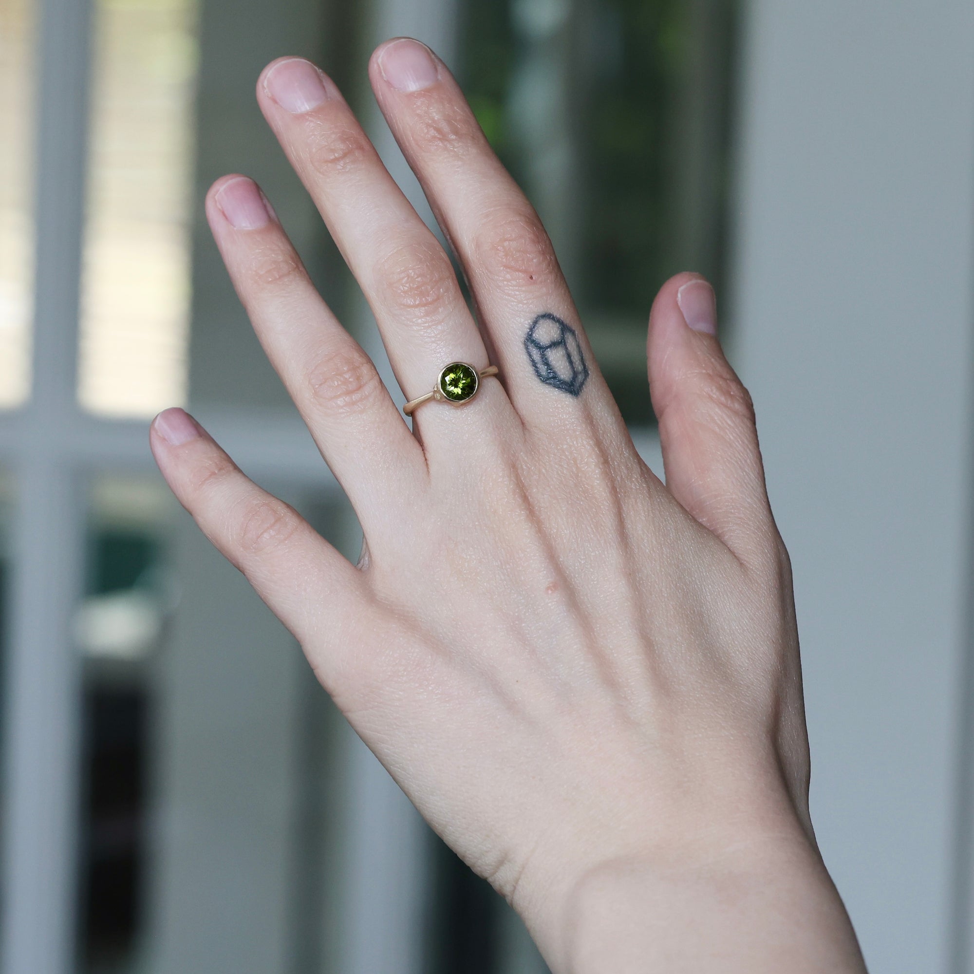 Hand wearing a ring with a green gemstone, finger tattoo, against a blurred background