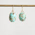 A pair of oval turquoise drop earrings with gold hooks and prong settings, hanging from a gold rod against a light gray background.