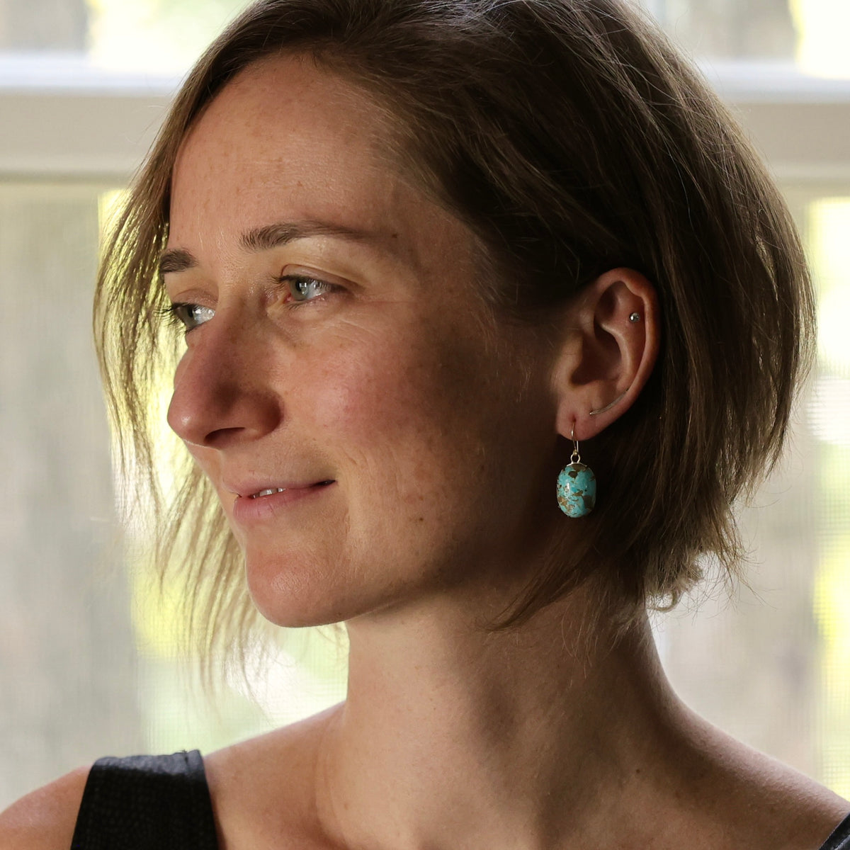 A woman in profile wearing a small turquoise drop earring, photographed indoors with soft natural light from a window behind her.