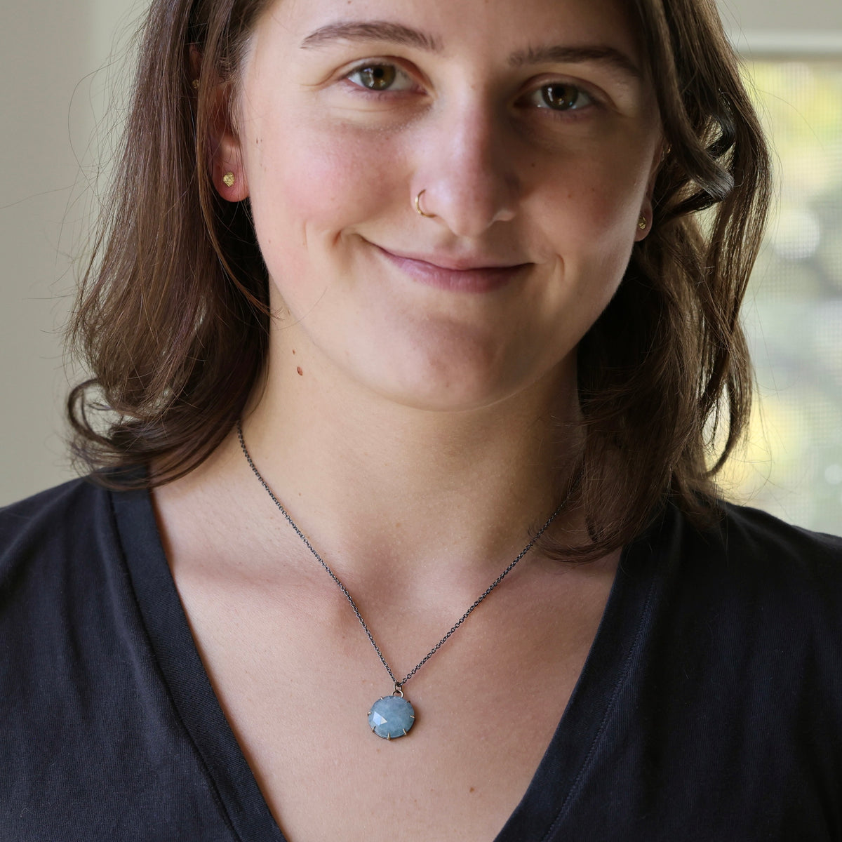 A woman with brown hair and a nose ring wearing a silver chain necklace with a round light blue stone pendant, photographed in a close-up portrait.