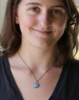A woman with brown hair and a nose ring wearing a silver chain necklace with a round light blue stone pendant, photographed in a close-up portrait.