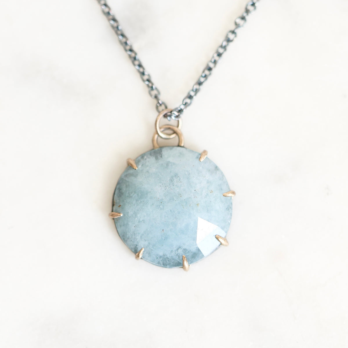 A round light blue gemstone pendant necklace with gold prong settings and a silver-toned chain, displayed on a light marble surface.
