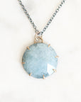 A round light blue gemstone pendant necklace with gold prong settings and a silver-toned chain, displayed on a light marble surface.