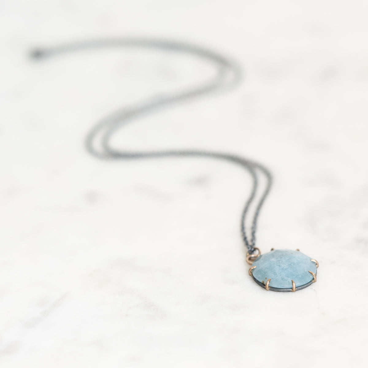 A delicate dark gray chain necklace with a round light blue gemstone pendant in a gold-toned bezel setting, resting on a white marble surface.