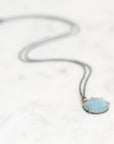 A delicate dark gray chain necklace with a round light blue gemstone pendant in a gold-toned bezel setting, resting on a white marble surface.