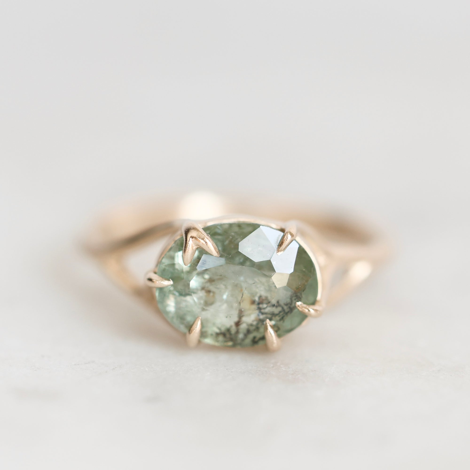 A close-up shot of a gold ring featuring an oval-cut light green gemstone with inclusions, set in a prong setting on a light gray background.