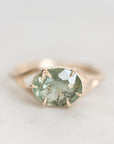 A close-up shot of a gold ring featuring an oval-cut light green gemstone with inclusions, set in a prong setting on a light gray background.
