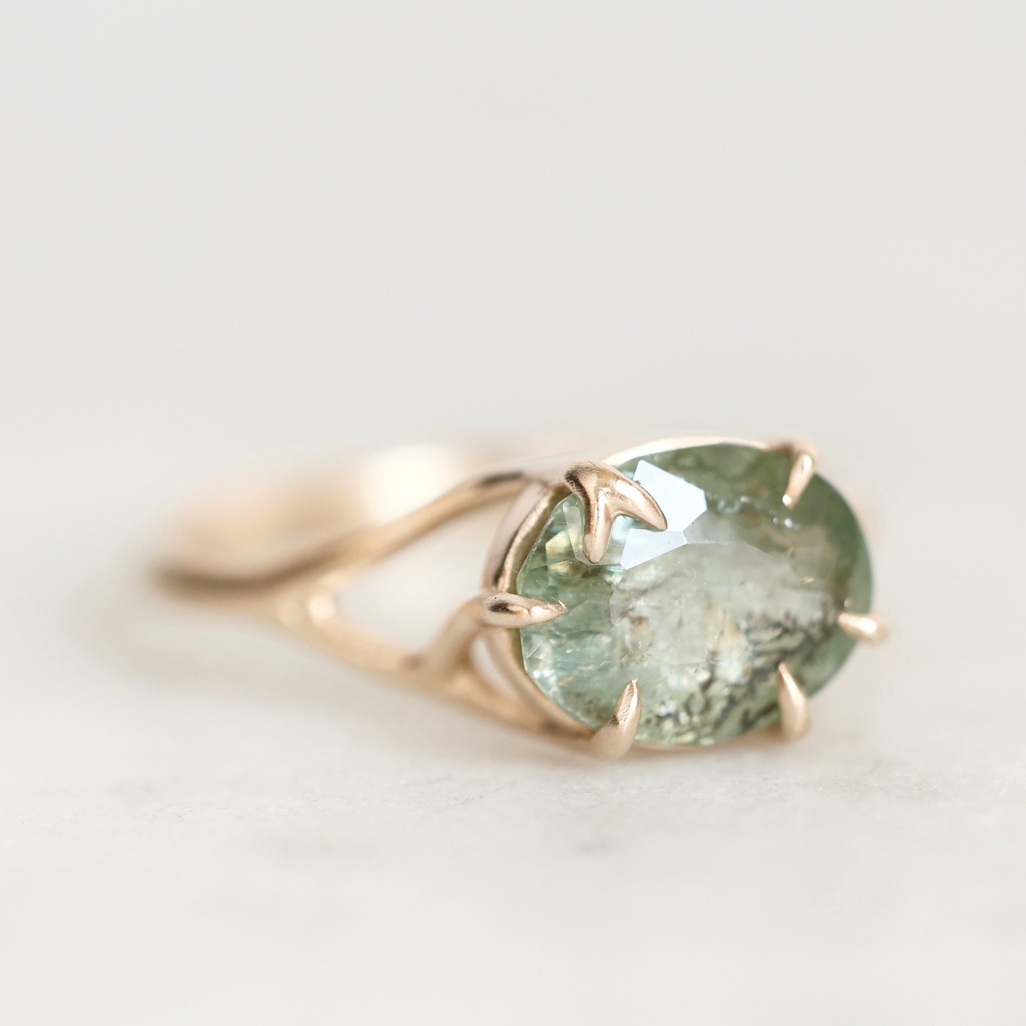 A gold ring featuring an oval-cut light green gemstone in a prong setting, shown on a soft, light gray background.