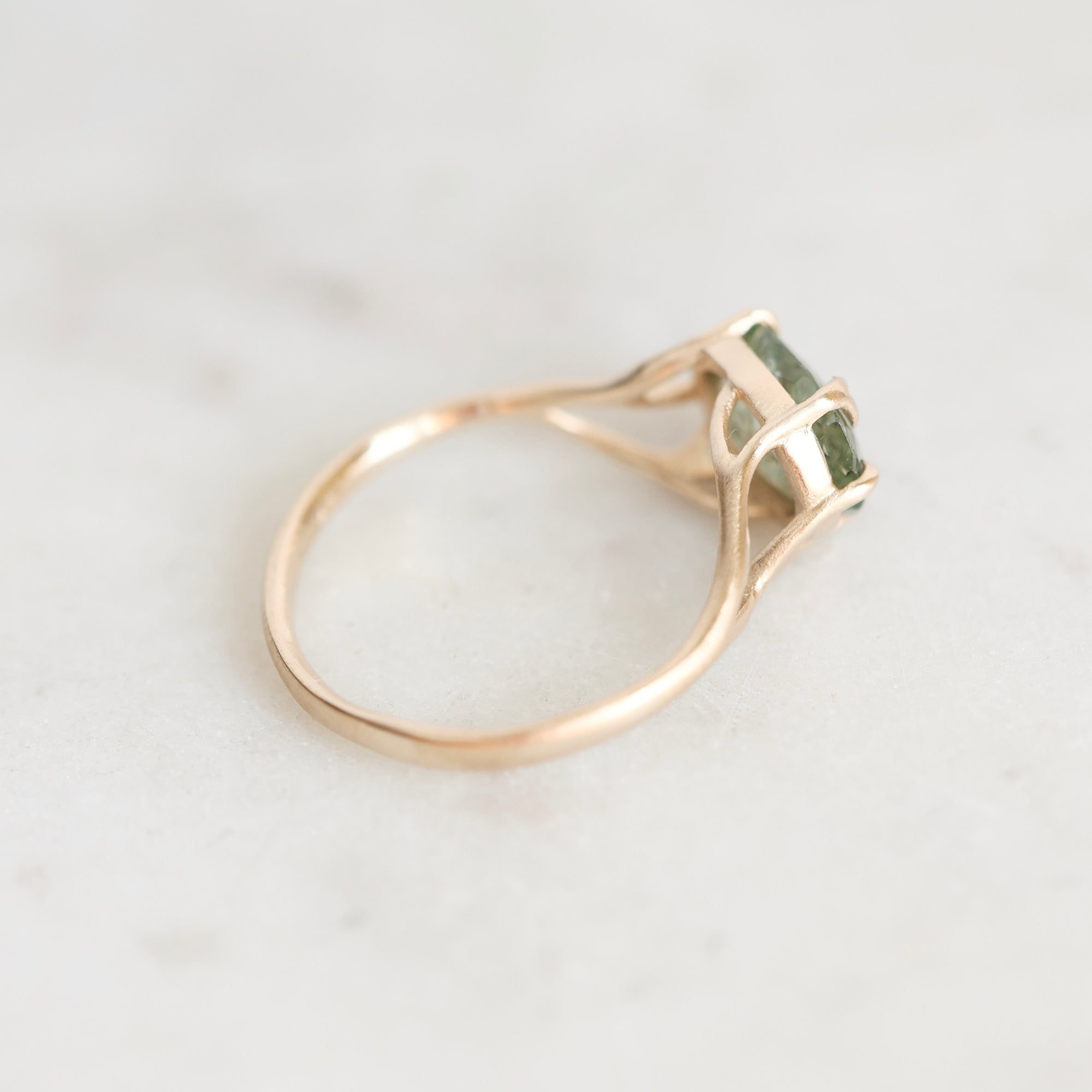 A rose gold ring with a square-cut green gemstone in a prong setting, resting on a light marble surface.