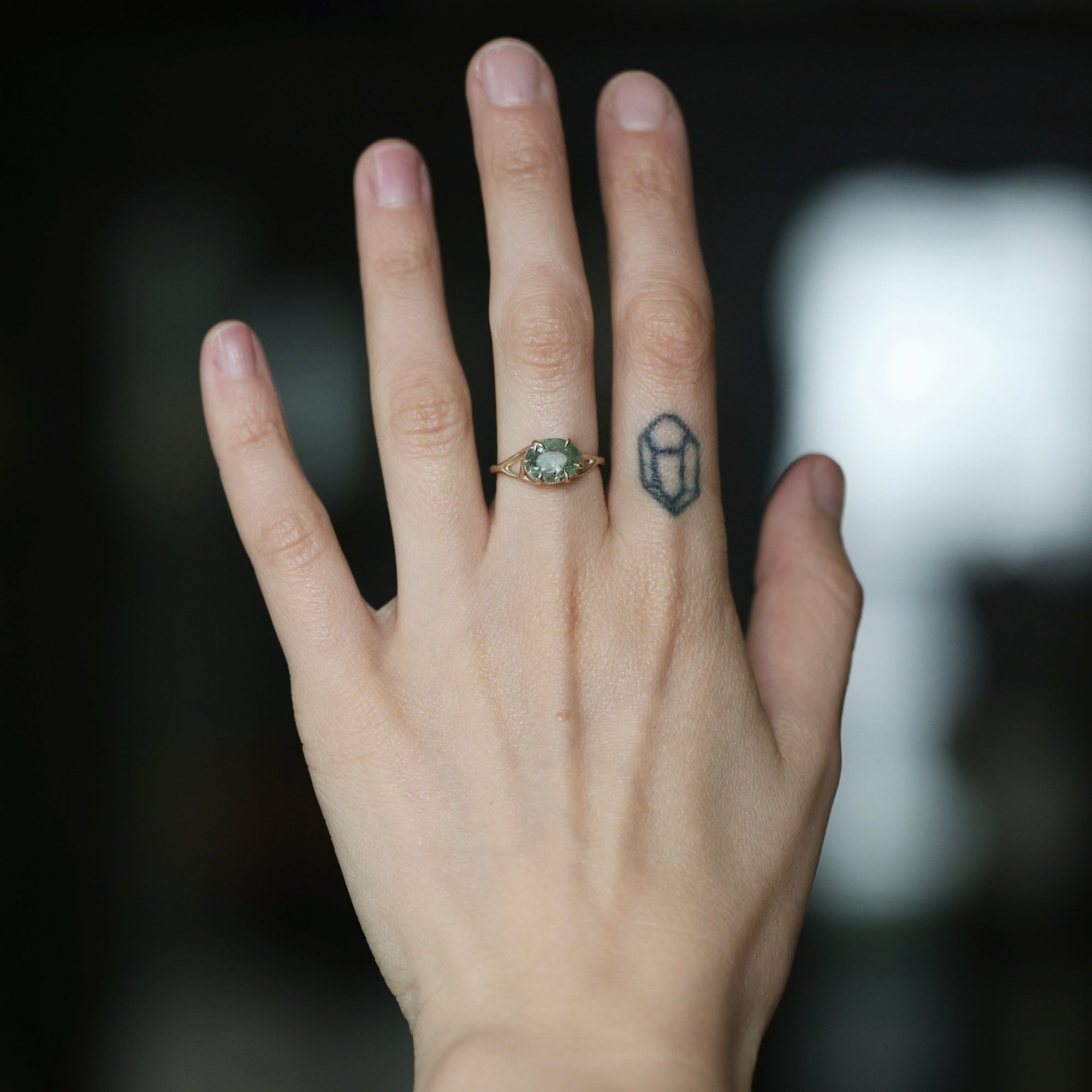 A close-up of a person's hand showing two rings: a small gold ring with a green oval stone on the ring finger, and a black geometric line tattoo on the middle finger. The background is dark and blurred.